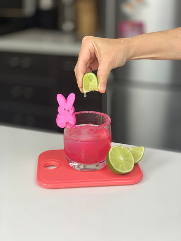 Hand squeezing lime over bright pink cocktail garnished with a pink marshmallow peep on a sugared rim.