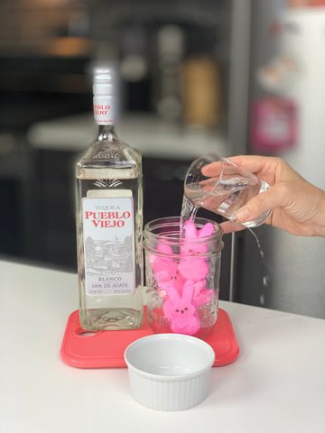 Tequila bottle labeled 'TEQUILA PUEBLO VIEJO BLANCO 100% DE AGAVE' beside a mason jar of pink Peeps while liquid is poured.