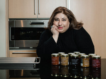 Woman leaning at kitchen counter behind jars labeled 'spiced raisin marmalata', 'brinjal caponata', 'tomato and white sultana chutney' LEBONMAGOT