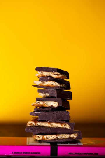 Stacked dark chocolate with pretzel layers on pink box; visible words: A WOMAN-OWNED BUSINESS, LEGALLYADDICTIVEFOODS.COM