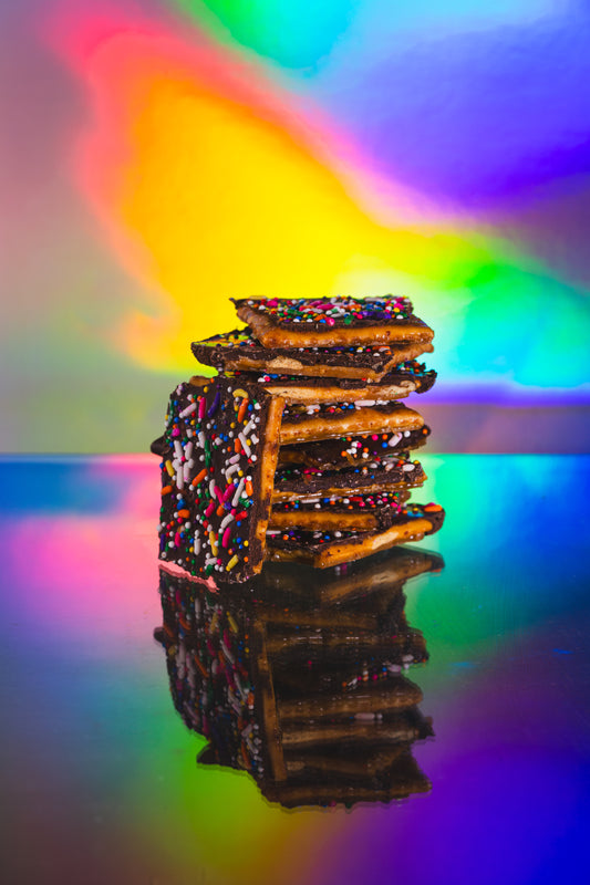 Stack of chocolate-covered pretzels with rainbow sprinkles reflected on a glossy surface