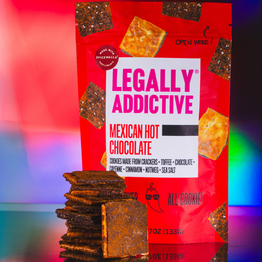 Red bag labeled LEGALLY ADDICTIVE MEXICAN HOT CHOCOLATE beside stacked chocolate squares and a blue sticker.